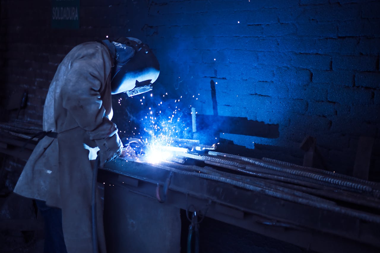 about-us Industrial worker welding metal indoors, emitting bright blue sparks and smoke.
